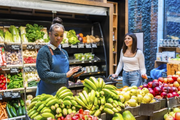 Grocery store employee using a digital tablet for inventory management of fresh fruits and vegetables while customer choosing products