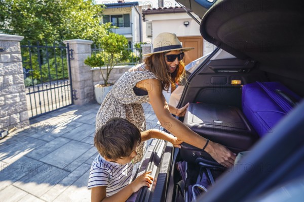 Mother and son preparing for a road trip, loading suitcases into the trunk of their car in front of their house