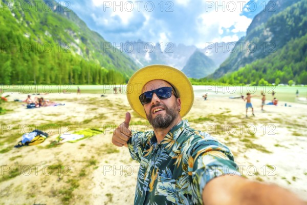 Tourist wearing sunglasses and straw hat gesturing thumbs up while taking a selfie at lago di landro with tourists enjoying summer in the dolomites, italy