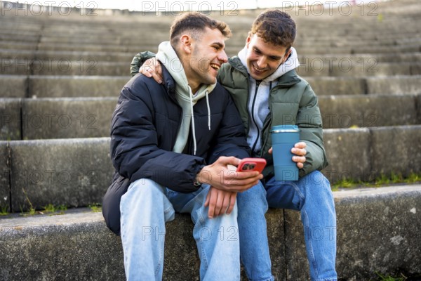 Two men are sitting on steps, enjoying a reusable coffee cup and sharing a laugh as they look at a smartphone together