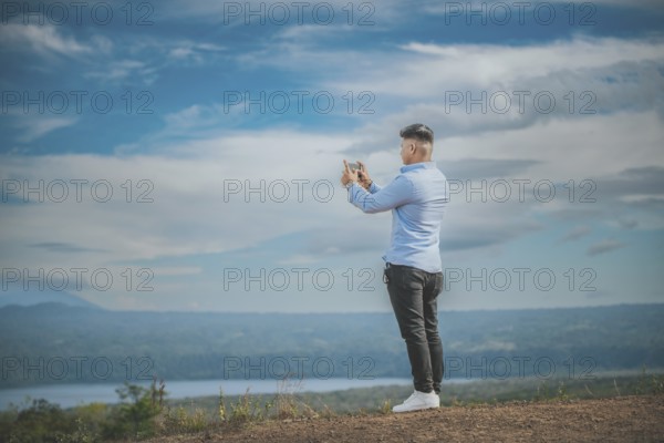 Young man taking photos with cell phone at a natural viewpoint. Tourist taking photos with phone at a volcanic viewpoint, Masaya, Nicaragua, Central America