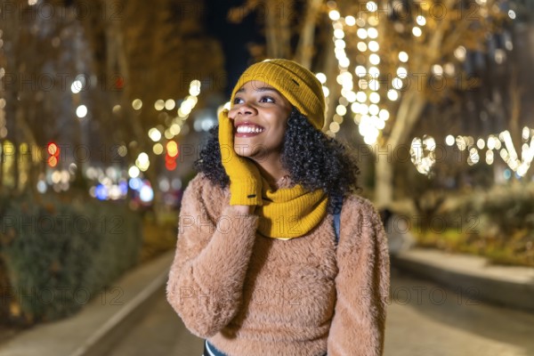 Smiling young woman wearing winter clothes touching her face while enjoying christmas lights in a city street at night