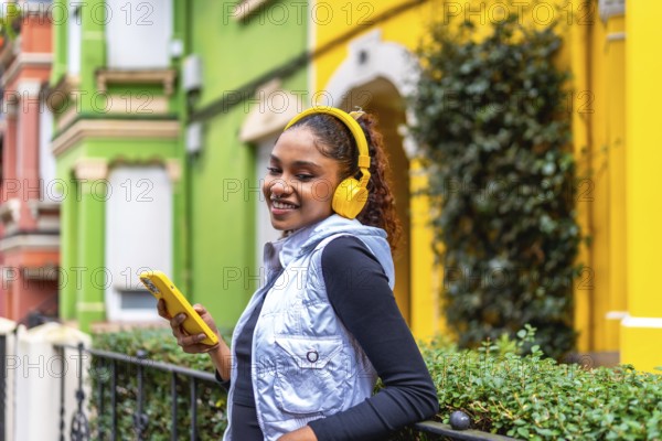 Happy african american woman using phone and listening to music in the city next to colorful houses