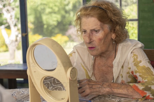Senior woman applying makeup while gazing at her reflection in a double mirror on a table, illuminated by bright natural light streaming through a window in the background