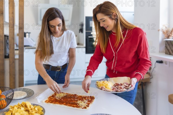Two women are happily adding cheese to a pizza base while preparing a meal together in a modern kitchen