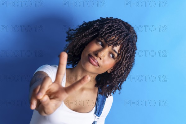 Young woman with curly hair making a victory sign with her fingers against a vibrant blue background, radiating confidence and positivity