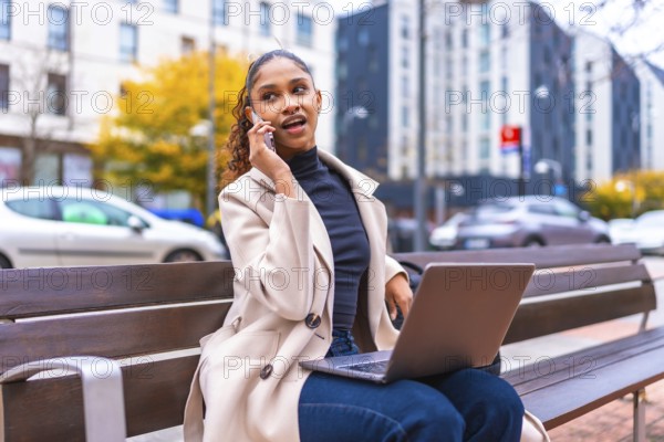 Busy young african american businesswoman using laptop talking to phone sitting on bench in the city