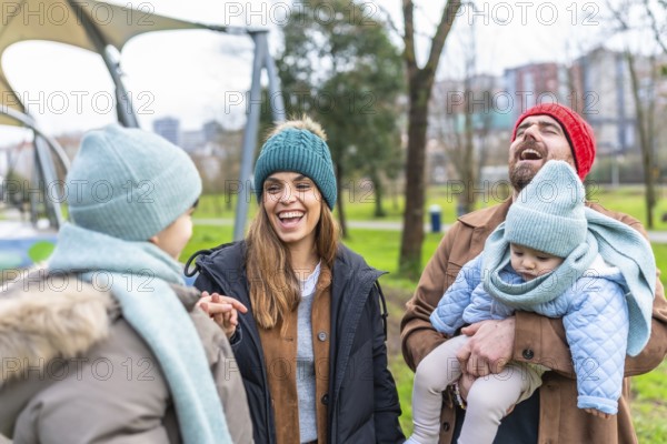 Happy parents holding their baby, laughing and talking with their other child in a park during winter