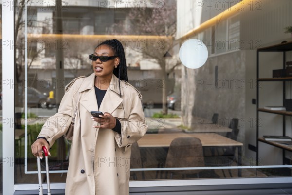 Stylish businesswoman using her smartphone while waiting outside a hotel lobby with her trolley suitcase, ready for her next business trip