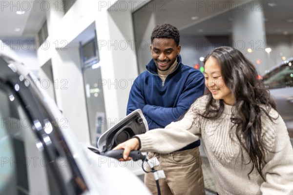 Happy multiethnic couple charging their new electric car at a dealership showroom, embracing a sustainable lifestyle and green energy solutions