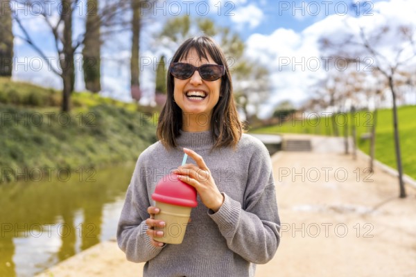 Young woman smiling and holding a reusable cup with a straw, enjoying a refreshing drink in a sunny park during springtime