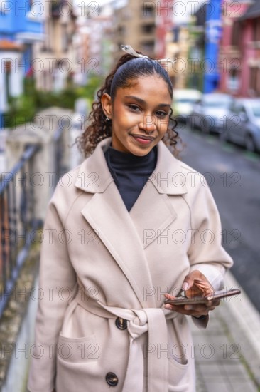 Vertical portrait of an elegant african american woman in tench coat using phone in the city