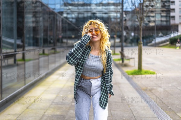 Stylish young woman with blond curly hair and sunglasses dances in a modern urban setting, enjoying the music and city vibes