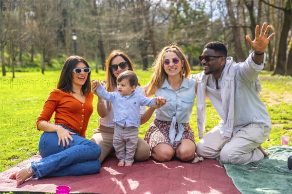 Group of cheerful multi ethnic friends with sunglasses enjoying picnic in a park with a baby boy waving