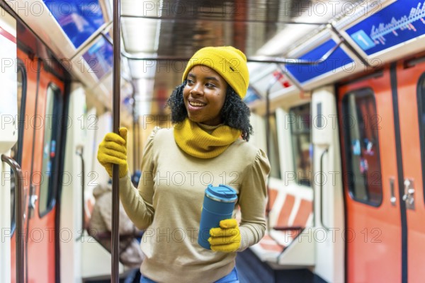 Smiling woman wearing yellow woolen hat and gloves holding a reusable mug while standing inside a subway car