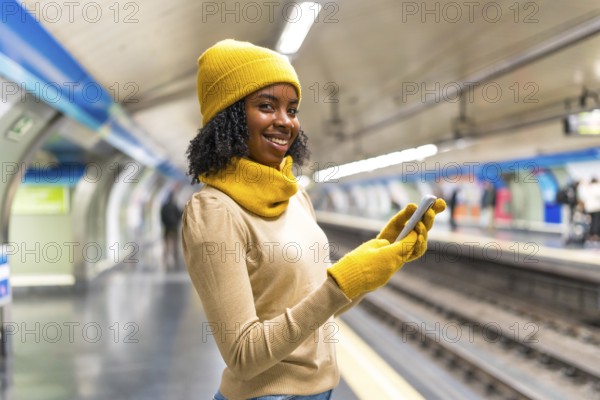 Young black woman wearing yellow winter clothes and gloves using her smartphone while waiting for subway train