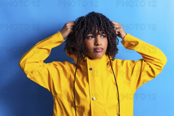 Young woman with curly hair wearing a yellow raincoat adjusting the hood while looking away on a vibrant blue background