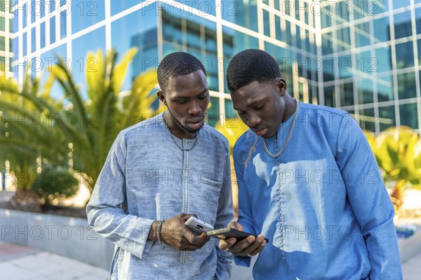 Two senegalese businessmen wearing traditional dashiki attire are using smartphones in front of a modern office building