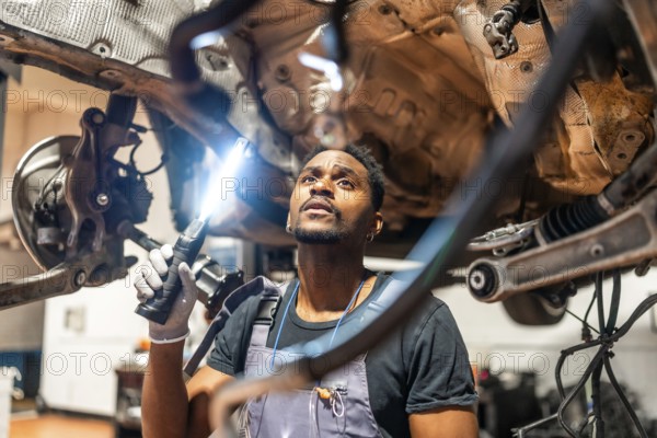 Young mechanic using flashlight, inspecting underside of car in a professional workshop, focused on maintenance and repair