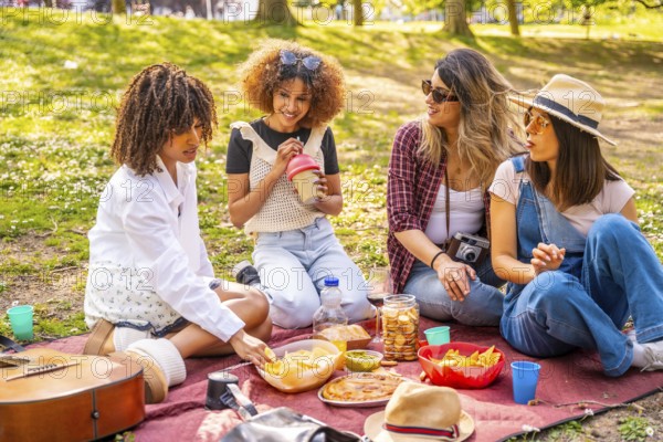 Four female friends are sitting on a blanket, eating snacks and drinking beverages during a picnic in a sunny urban park