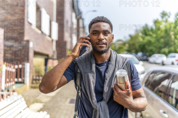 Young african man talking to the mobile commuting to work carrying a hot coffee thermos along a residential district