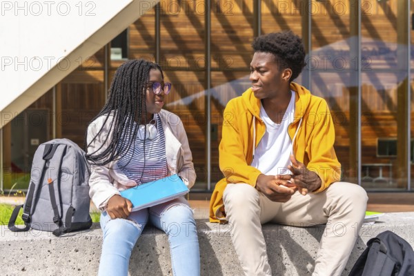 Two young African american students chatting relaxed outside the university sitting together under the sun