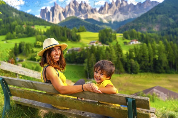 Mother and son sitting on a bench, admiring the breathtaking landscape of the italian dolomites near santa maddalena church in val di funes