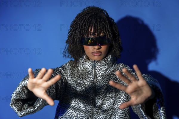 Young woman wearing futuristic visor and patterned jacket, gesturing with hands on blue background, immersed in metaverse gaming experience