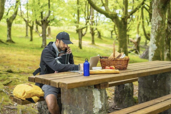 A young Caucasian father teleworking sitting at a picnic table with his computer and talking on the phone, with the boy in his backpack. Summer lifestyle