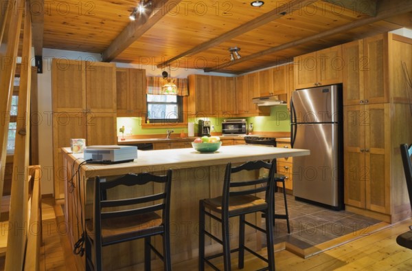 Wooden island with ceramic tile countertop, bar stools and shaker style cabinets in kitchen with lights on inside Canadiana style log cabin home, Quebec, Canada, North America