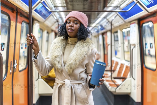 Stylish young black woman wearing winter coat and pink beanie holding reusable coffee cup while commuting on subway train