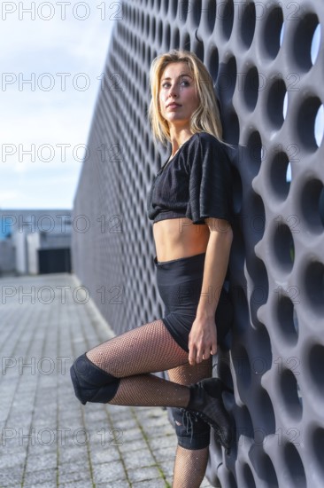 Young urban dancer striking a pose in high heels, knee pads, and fishnet stockings, exuding confidence and style against a city backdrop