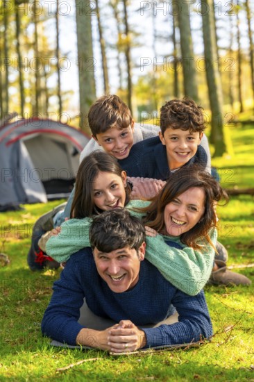 Vertical photo of a caucasian cheerful family laughing while lying down together at forest next to camping tent
