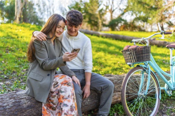 Smiling young couple sitting on a log in a park, sharing a moment on a smartphone, with a vintage bicycle nearby on a sunny day