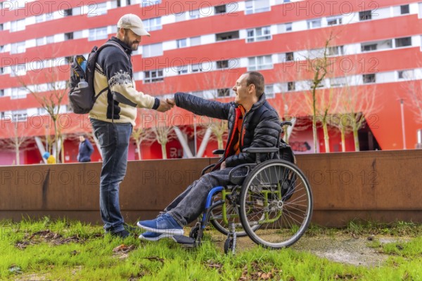 Tourist with backpack shaking hands with a disabled man in a wheelchair in a city park, with a red building in the background