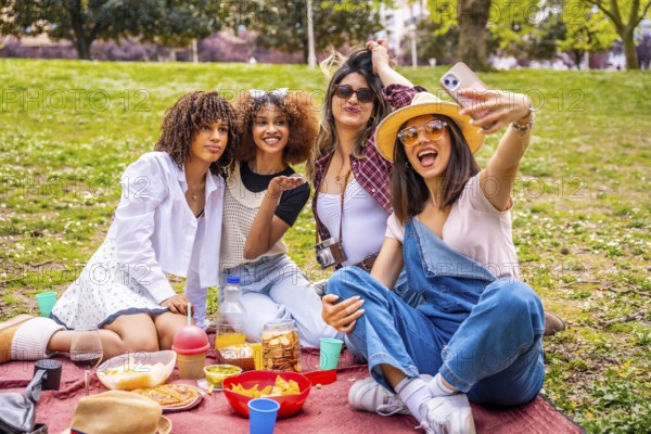 Four cheerful young women enjoying a sunny picnic in the park, capturing joyful moments together while taking selfies with a smartphone