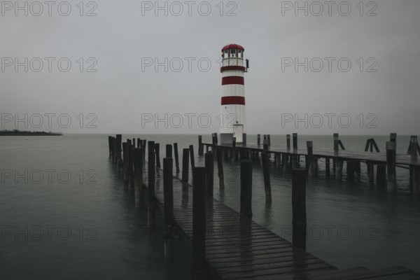 A lighthouse at the end of a wooden jetty under a grey sky over calm water, Podersdorf, Burgenland, Austria, Europe