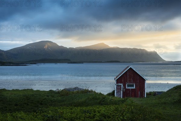 A red wooden hut on the sandy beach of Ramberg (Rambergstranda), with the sea behind it. At night at the time of the midnight sun. Clouds in the sky, the night sun shining from the side, mountain peaks in the sunlight. Early summer. Ramberg, Flakstadoya, Lofoten, Norway, Europe