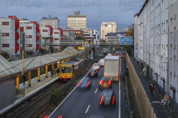 Essen, North Rhine-Westphalia, Germany, motorway A40 in the city centre, exit Essen Zentrum, during dusk. Underground line U18 on the way to Berliner Platz stops at Savignystrasse underground station. On the right, residential buildings behind a noise barrier. A LANUV air monitoring station, Essen Kruppstrasse 117 (EKRU2), is located on the wall on the right under the lantern. Residents are affected by noise and exhaust fumes. Building with Postbank, Eon and Evonik lettering at the back, Europe