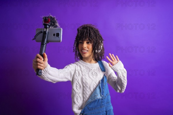 Young latin american content creator waving and recording video with smartphone and external microphone on purple background