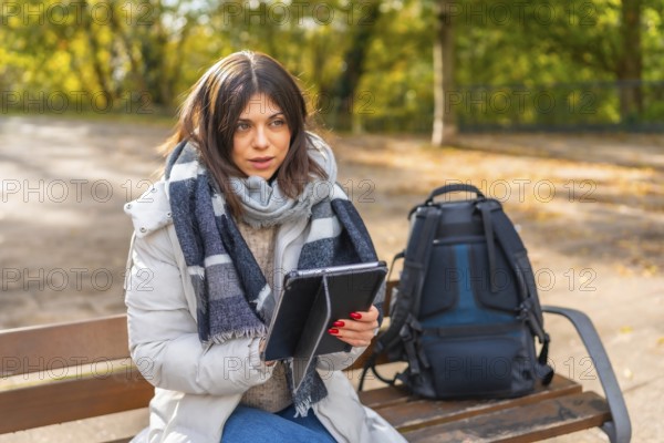 Woman with warm clothes working using digital tablet sitting on a park bench during sunny day of autumn