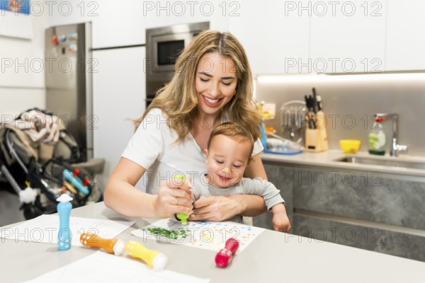 Happy mother and baby boy enjoying creative time together, painting with colorful markers in a bright, modern kitchen
