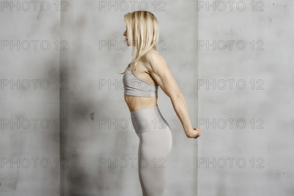 An athletically built woman stands in profile against the background of a concrete wall and stretches the muscles of her arms and back