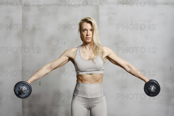 A middle-aged woman spreads her arms with dumbbells to the sides in gym