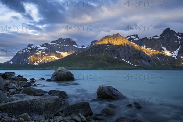 Landscape with sea and mountains on the Lofoten Islands, view over the fjord Flakstadpollen to mountains. Rocks in the foreground. At night at the time of the midnight sun, some clouds in the sky, sun spots on the mountains. Long exposure. Early summer. Flakstadoya, Lofoten, Norway, Europe