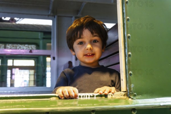 A young boy is sitting in a train car with his hands on the window. He is smiling and looking out the window