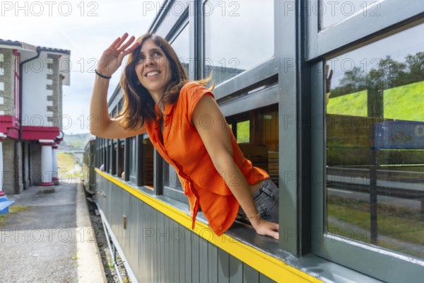 A woman in an orange shirt is looking out the window of a train. She is smiling and waving at the camera