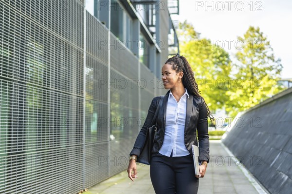 Horizontal photo with copy space of a modern latin businesswoman walking out the block office carrying laptop in a sunny day