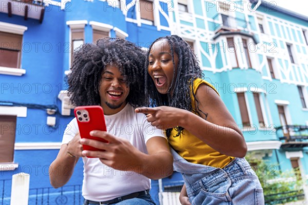 African friends pointing to a mobile with surprised expression standing next to blue colorful houses in the city