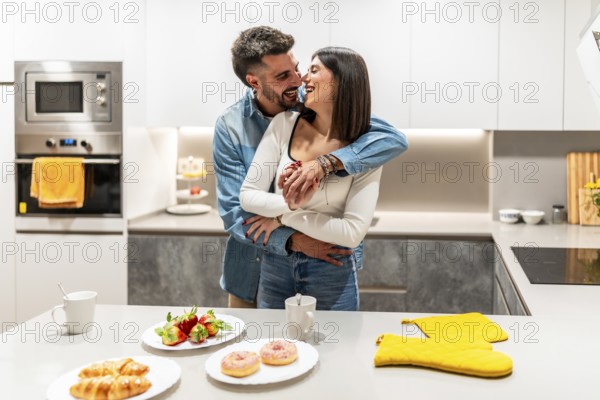 Young loving couple hugging and laughing in contemporary kitchen, with croissants, strawberries, and donuts on the counter, enjoying a romantic breakfast